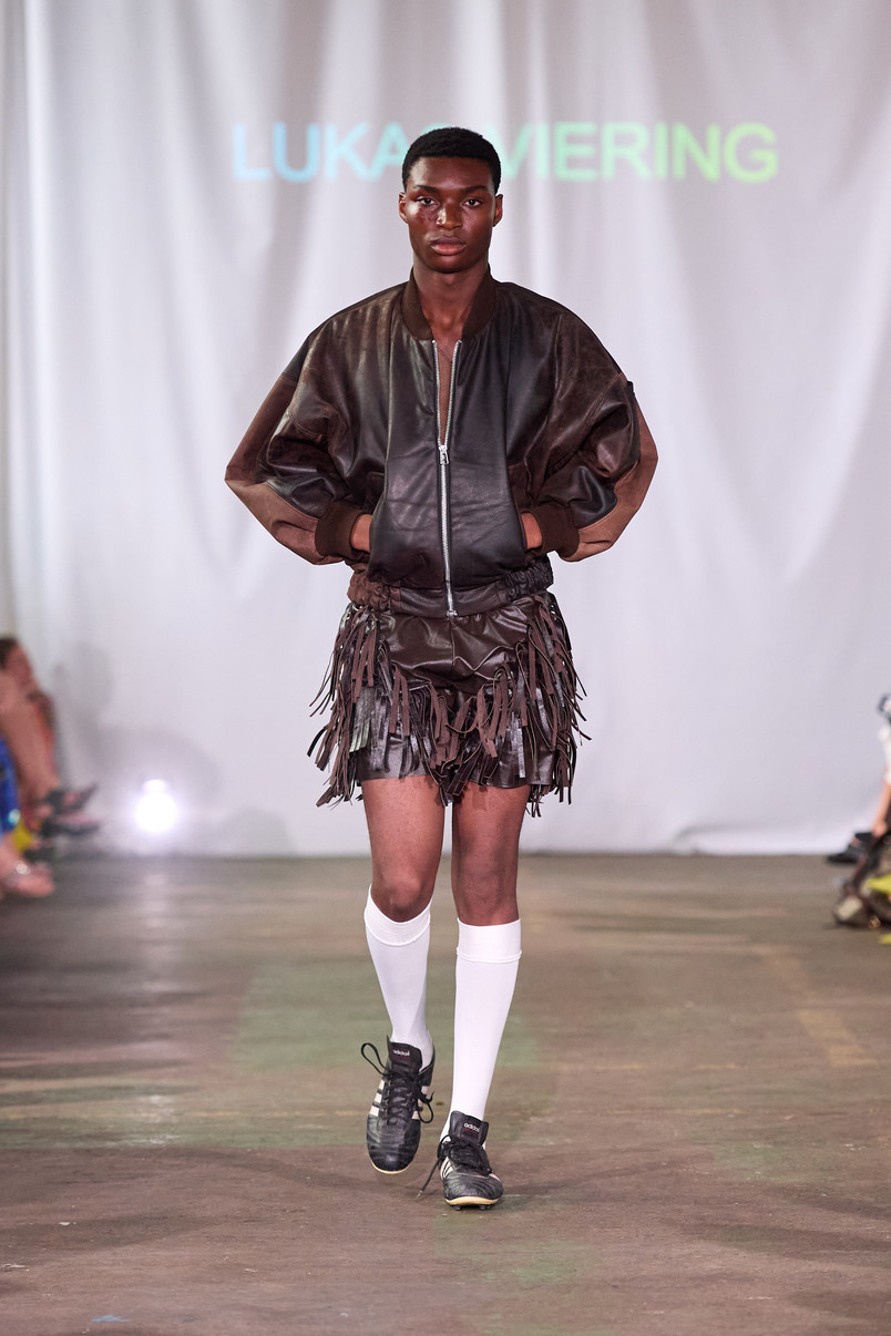 Model on a runway wearing a dark leather bomber jacket, fringed skirt, white knee-high socks, and black sneakers.