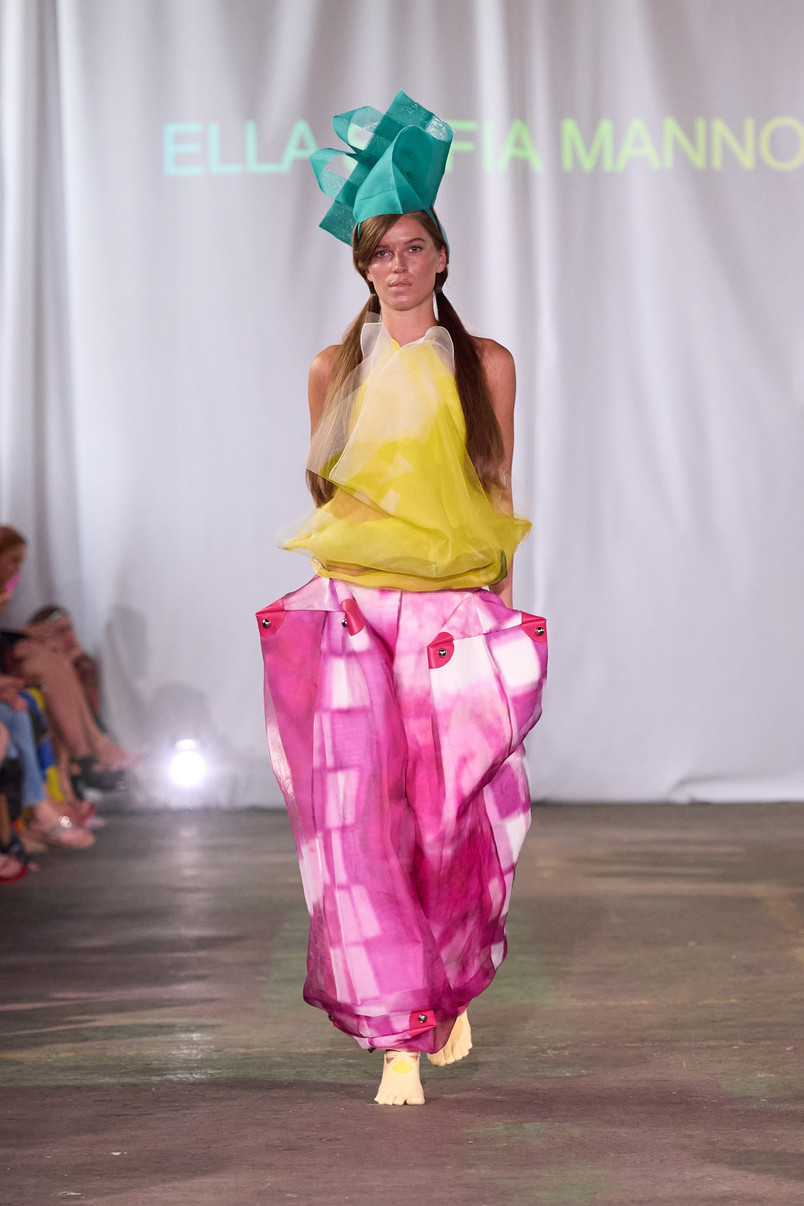 Model on a runway wearing a sheer yellow top, voluminous pink trousers, and a large turquoise headpiece.