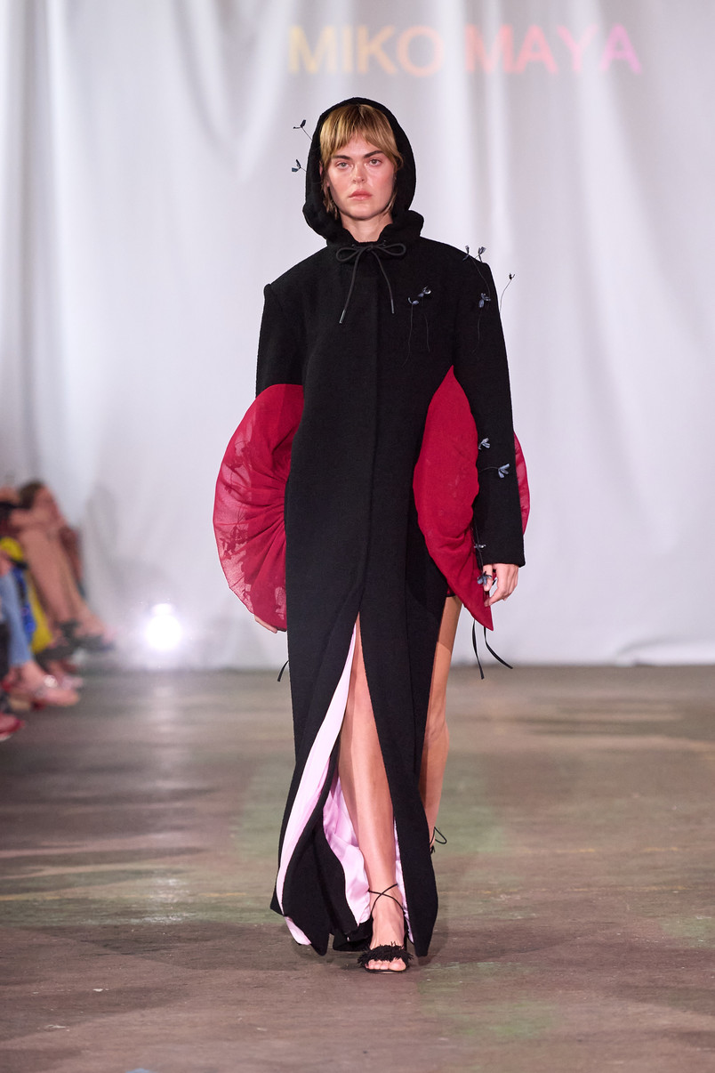 Model on a runway wearing a long black hooded dress with sculptural red side panels.