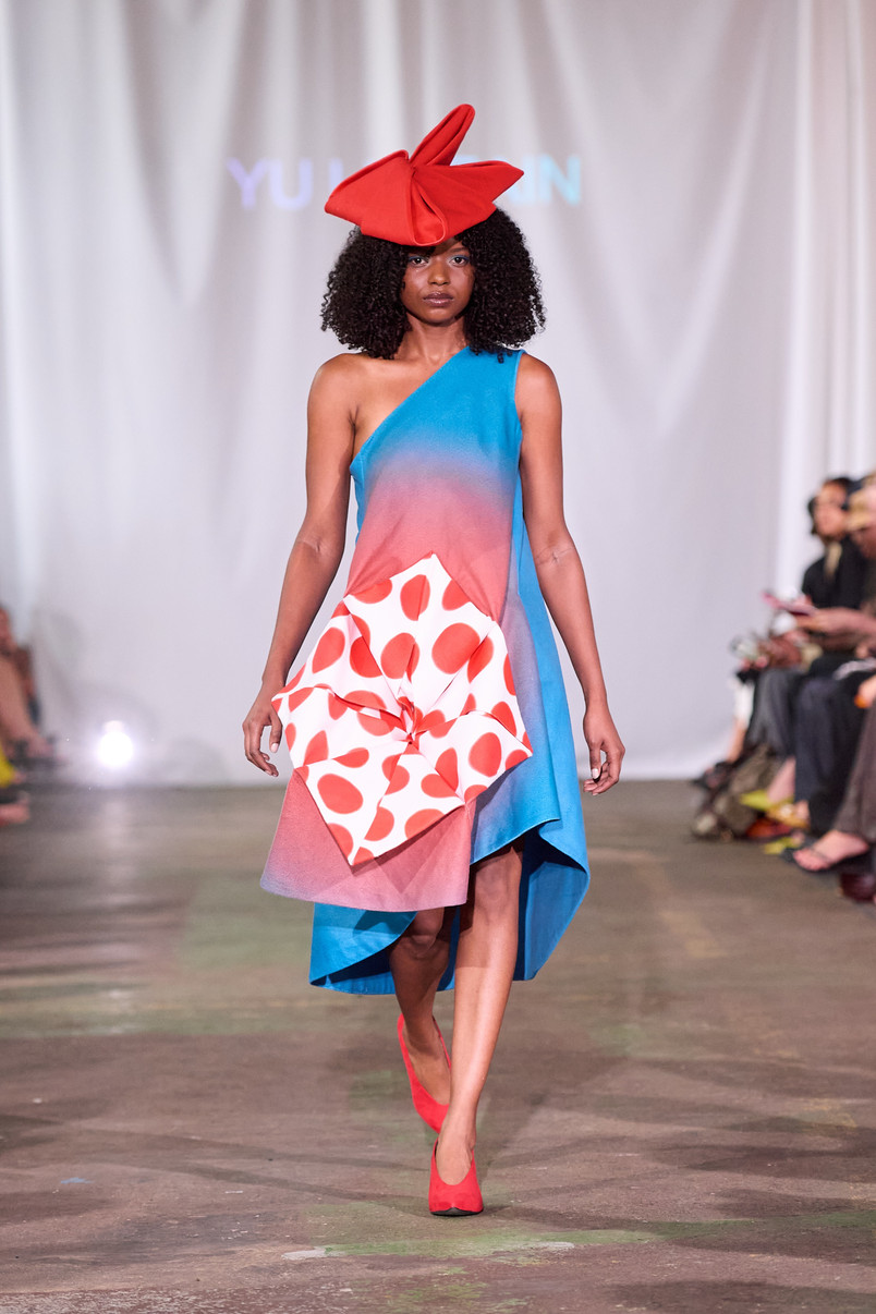 Model on a runway wearing an asymmetrical blue and pink dress with a sculptural red polka-dot panel and a large red hat.