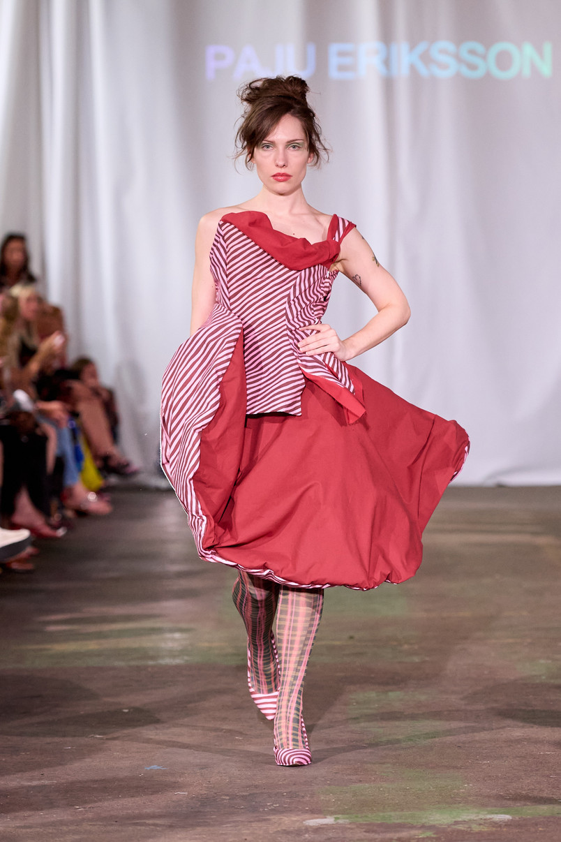 Model on a runway wearing a sculptural red dress with striped panels and patterned tights.