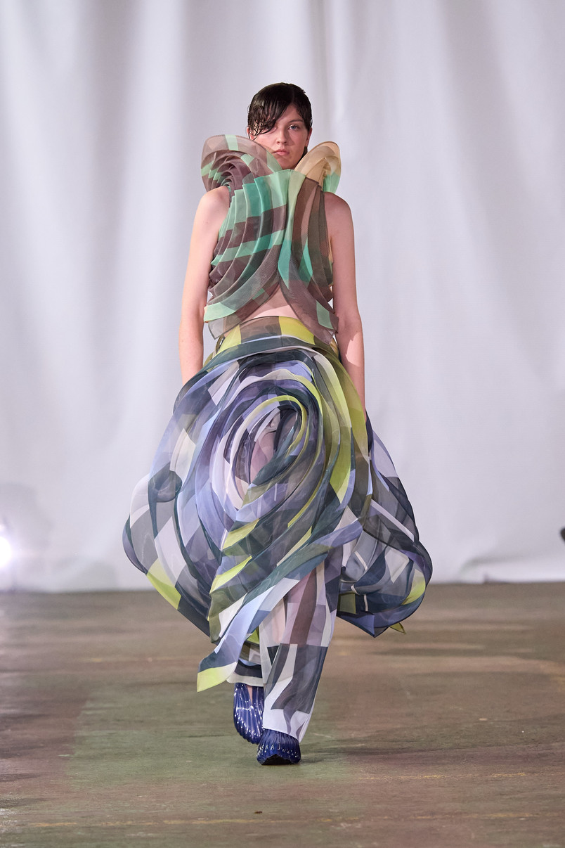 Model on a runway wearing a sculptural dress of sheer layered green, blue, and gray fabrics with flowing forms.