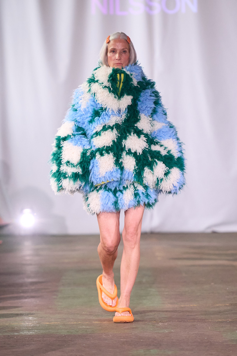 Model on a runway wearing a voluminous shaggy jacket in light blue, green, and white with orange sandals.