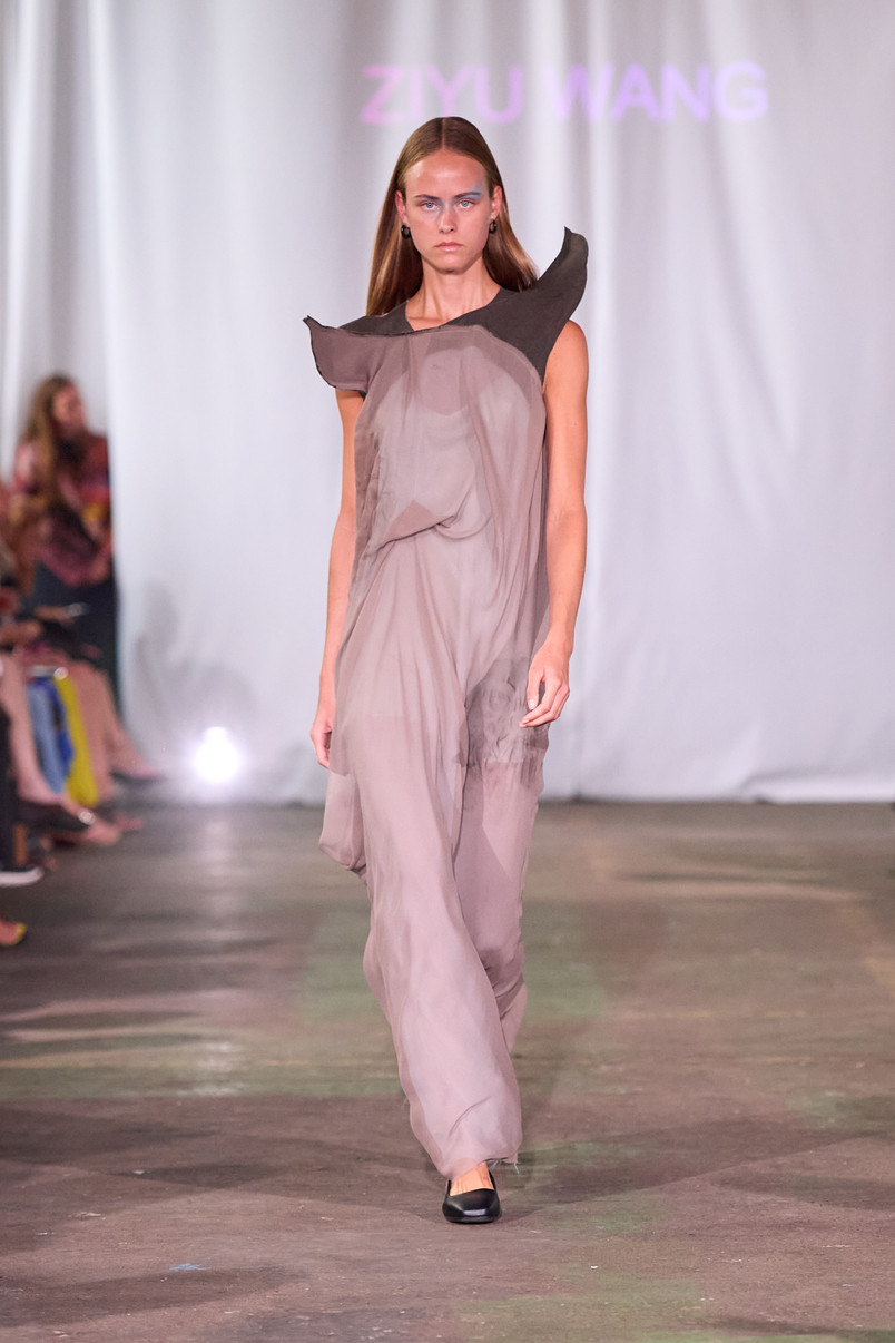 Model on a runway wearing a draped gray dress with sculptural black shoulder details.