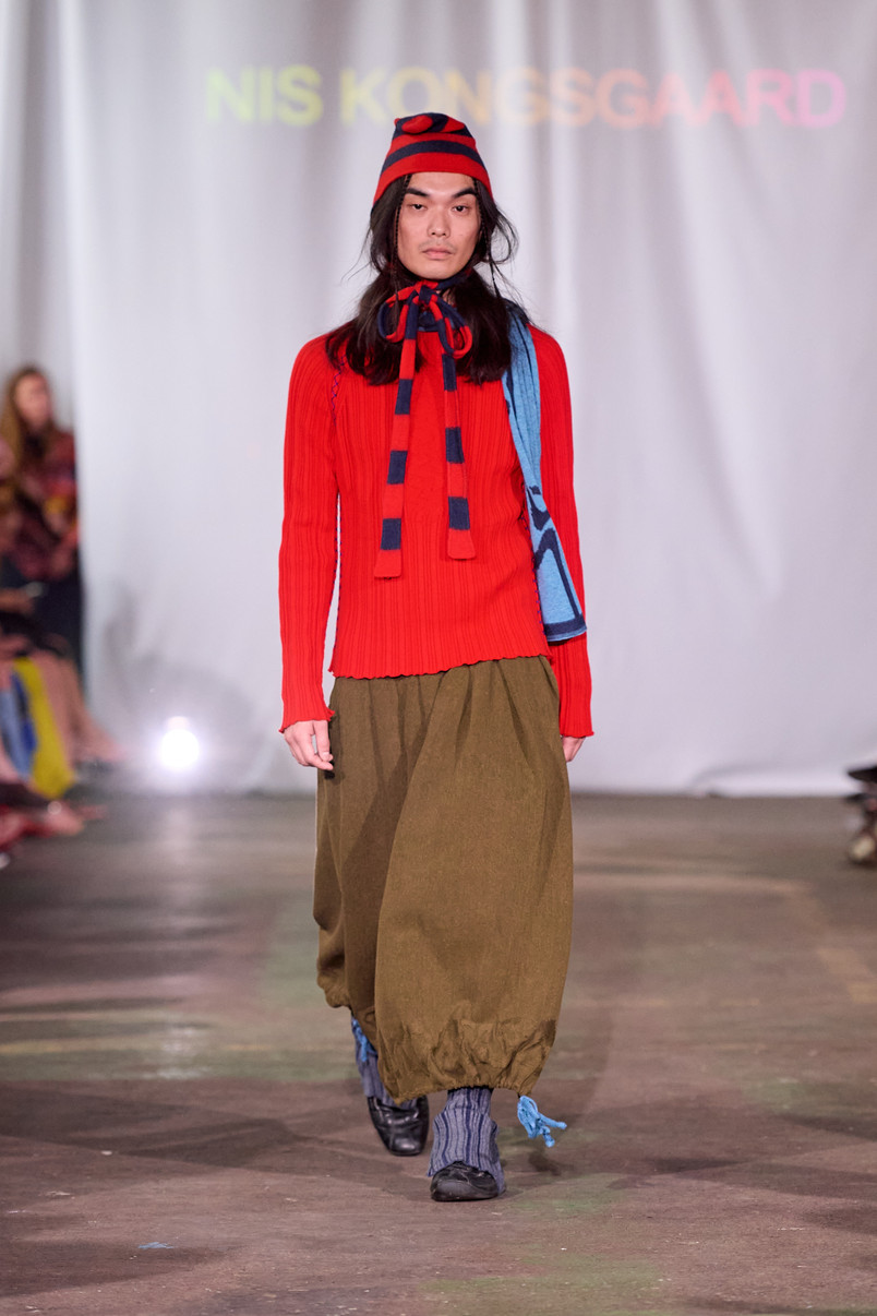 Model on a runway wearing a red ribbed sweater, a red and navy striped hat and scarf, and olive green balloon trousers.