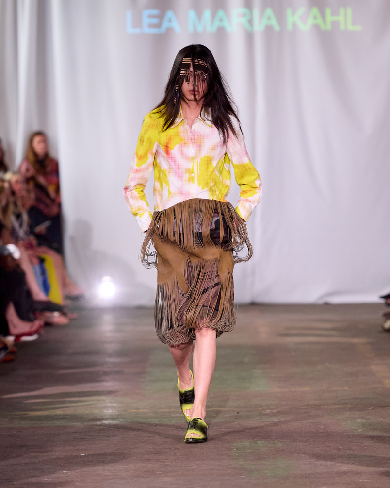 Model on a runway wearing a yellow and pink patterned shirt, a fringed skirt, and a beaded face covering.