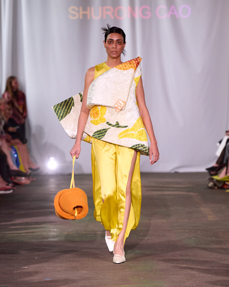 Model on a runway wearing a sculptural citrus-print top, yellow trousers, and a round orange handbag.