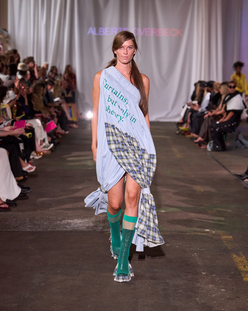 Model on a runway wearing a light blue draped dress with printed text, plaid details, and turquoise knee-high socks.