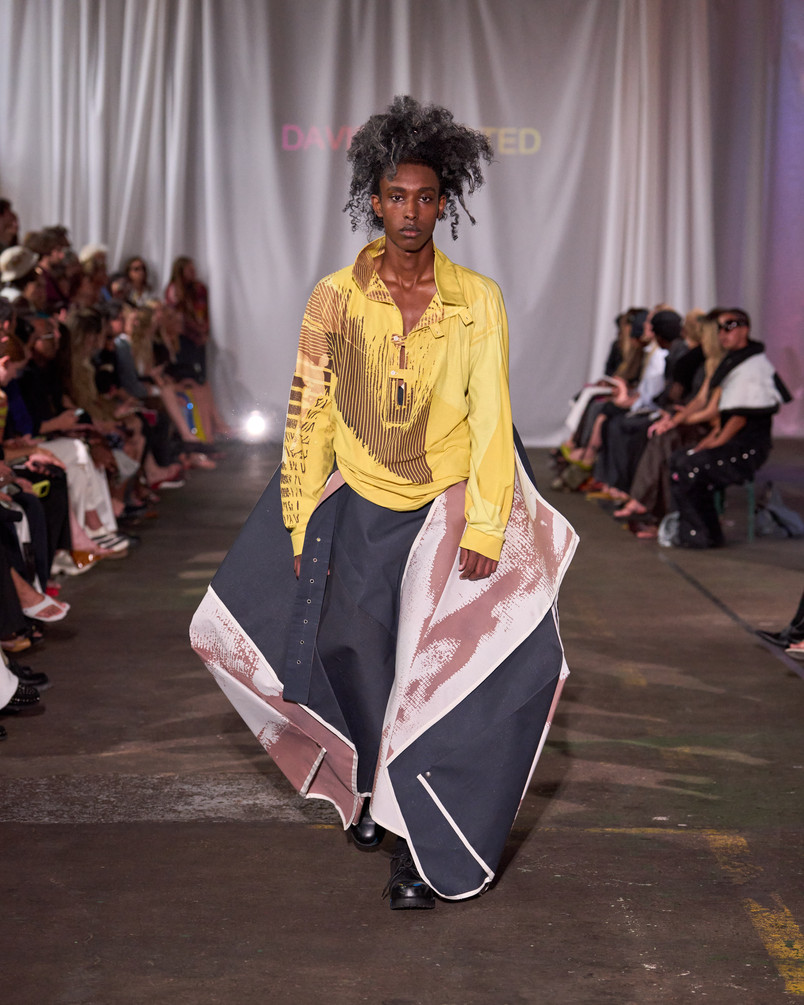 Model on a runway wearing a yellow patterned shirt and a voluminous skirt with graphic panels.