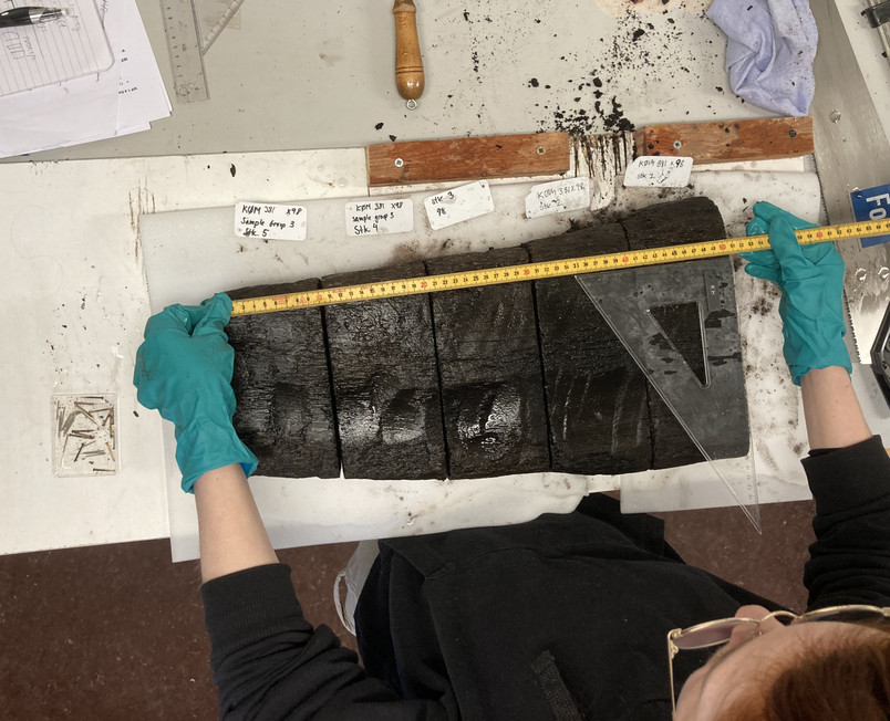 Gloved person measures sections of dark, aged wood on a workbench.