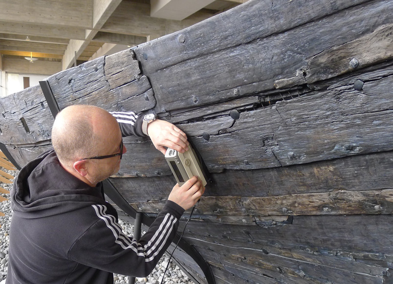 Person examines a large, aged wooden structure with a handheld measuring instrument.