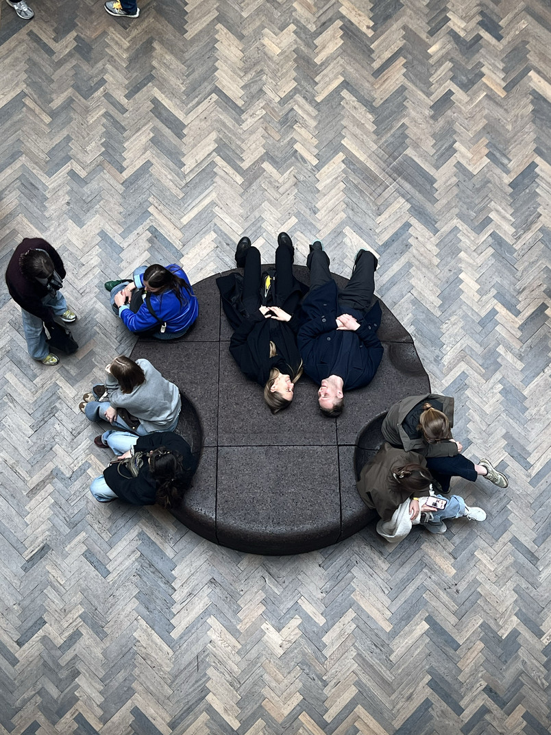 People are sitting and lying on a circular bench surrounded by herringbone-patterned wooden flooring.