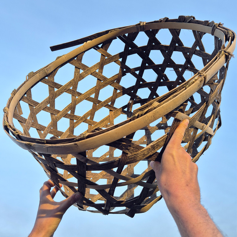 Two hands are holding up a large woven basket made of wooden strips against a blue sky.