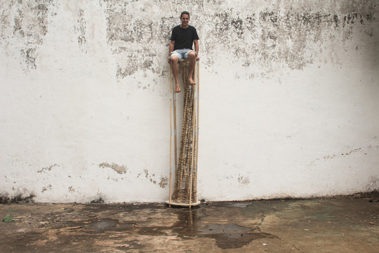 Person seated on a tall, narrow structure made of natural materials in front of a wal