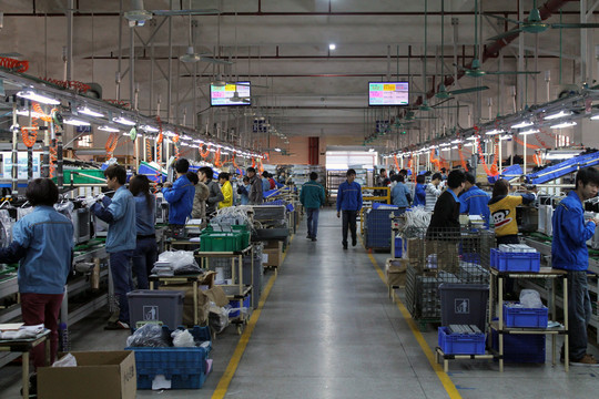 Workers on an assembly line in a large electronics factory with machines and components.