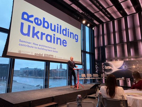 A speaker is standing on a stage in front of a large screen displaying the text ‘Rebuilding Ukraine’ during a seminar.