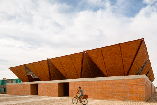 Modern brick building with a striking folded roof; cyclist rides past.