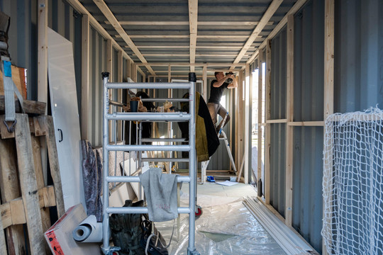 Person working on a ladder inside a container with timber framing under construction.