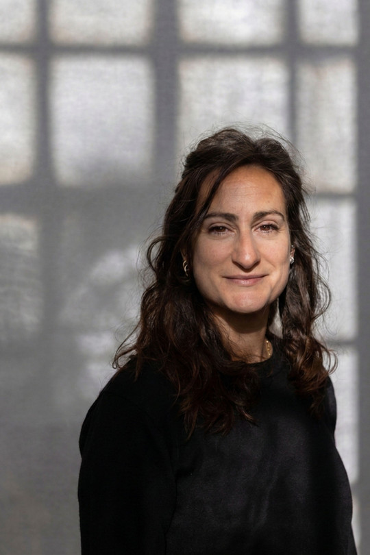 Picture of Roberta with shoulder-length brown hair and fair skin, wearing a light-colored t-shirt, looking directly at the camera with a neutral expression against a soft, light background.