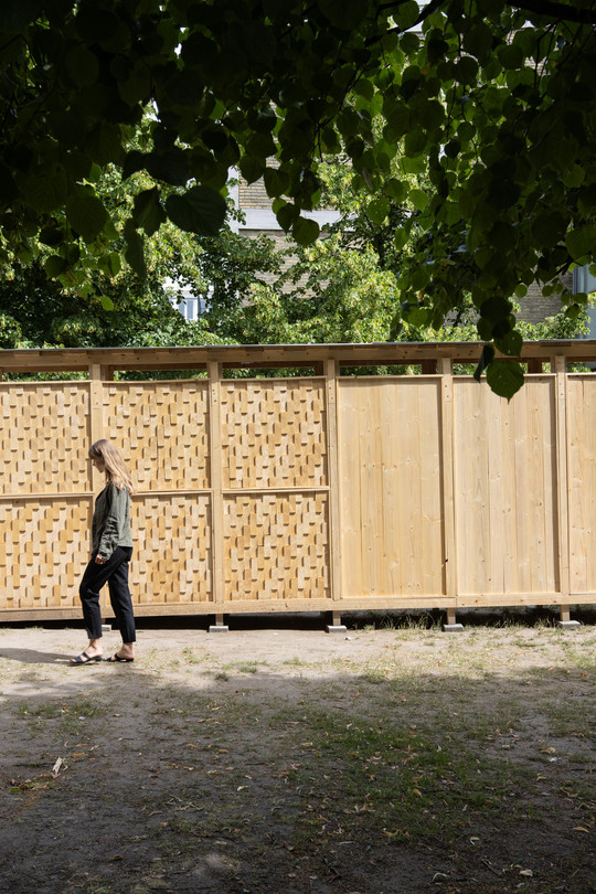 Woman walking past a wooden structure with smooth and woven panels beneath green trees.