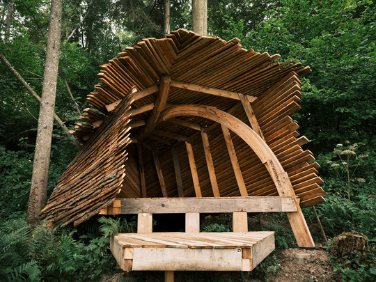 Sculptural shelter made of overlapping timber boards, with curved supporting beams and a platform in the forest.
