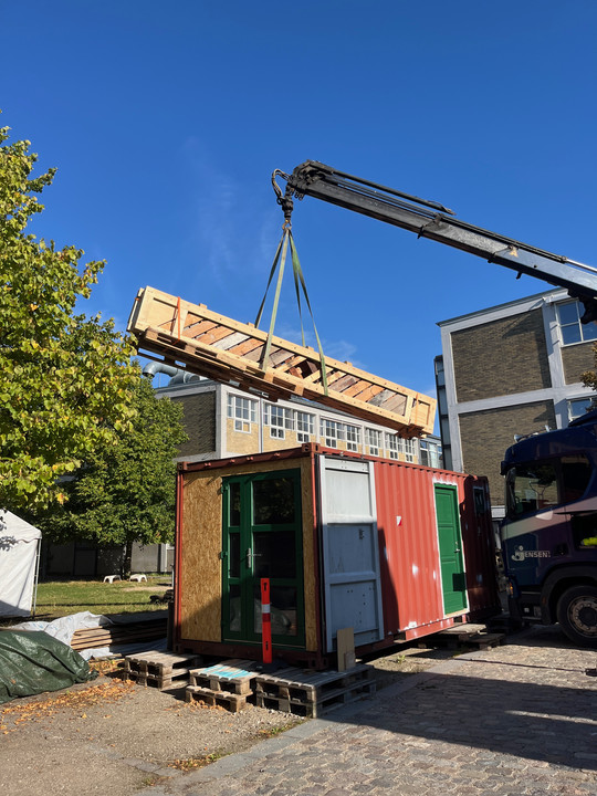 A crane lifts a long timber lattice structure above a red container next to an industrial building.