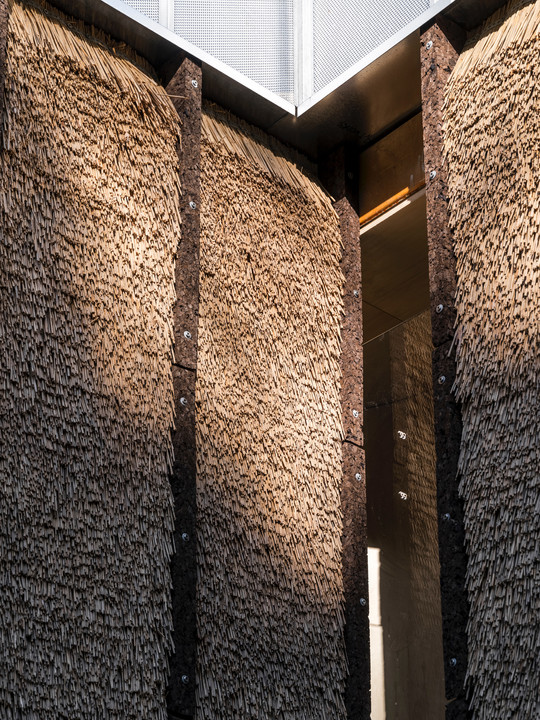 Façade detail with tightly packed bundles of thatching straw secured between dark vertical panels beneath metal cladding.