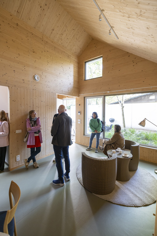 Several people are gathered in a bright communal space with timber-clad walls, a high ceiling and large windows.