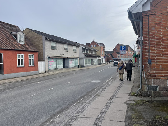 Two people walk along a quiet urban street lined with older brick buildings and vacant shop premises.