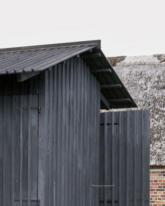Dark grey timber building with a pitched roof and double doors in front of a thatched house.