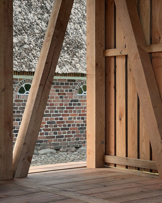 Interior timber structure with diagonal posts and plank flooring in front of an old brick building.