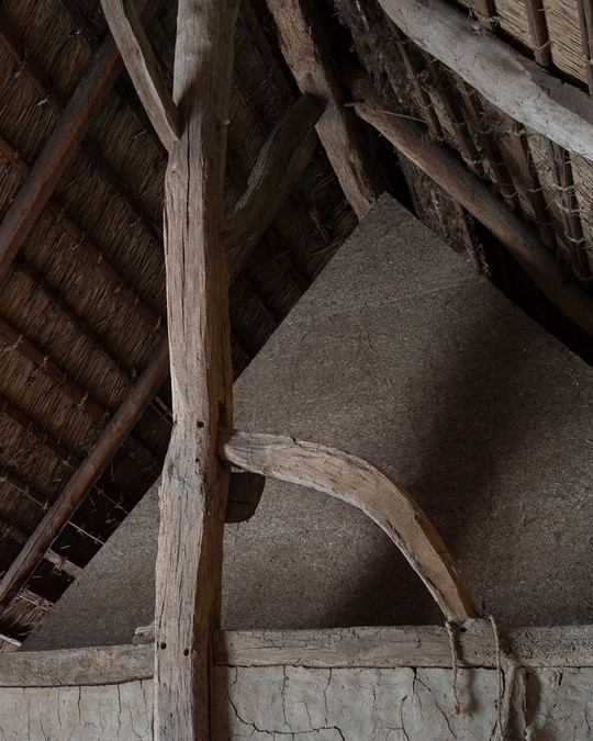 Old roof structure with substantial, curved timber beams beneath a thatched roof.