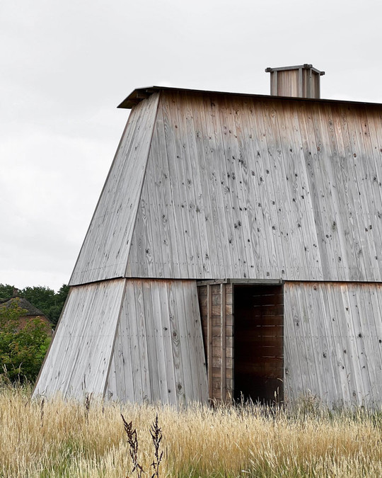 Tall, angular timber building with a grey façade, an open door, and tall grass around it.