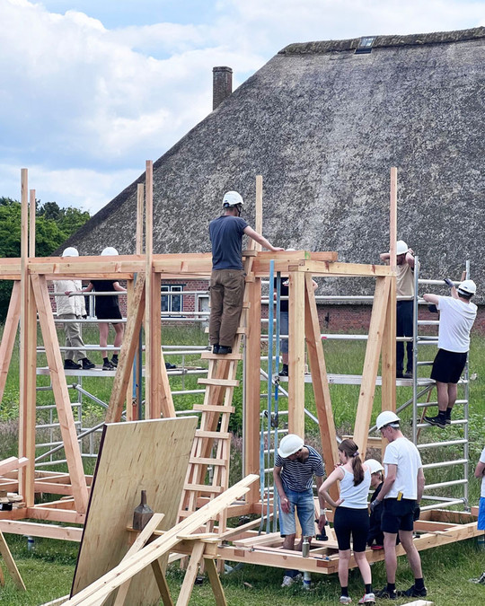 A group of people wearing safety helmets are building a timber structure on scaffolding in front of a thatched house.