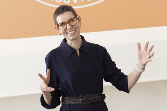 : Sofie Beier smiler og gestikulerer med begge hænder foran en orange og hvid baggrund.