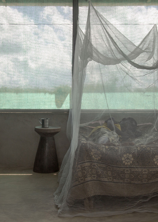 Bed with a patterned bedspread under a white mosquito net by a large mesh-covered window.