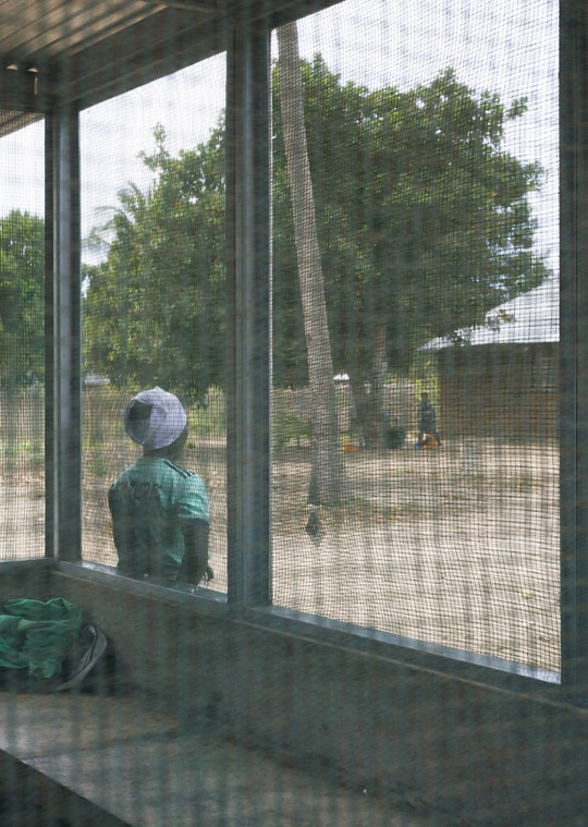 Person wearing a green jumper and a white hat stands outside a building with large mesh-covered windows.