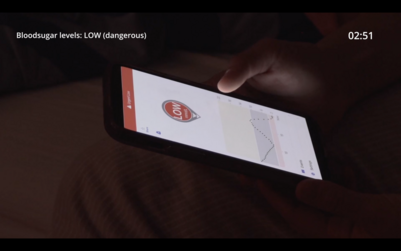 A person is holding a smartphone displaying a health app with a warning indicating "LOW" blood sugar levels, marked as dangerous. The screen shows a graph with fluctuating data points. The time on the screen reads 02:51, and a message at the top of the image states "Blood sugar levels: LOW (dangerous)."