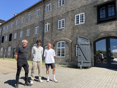 Tre mænd står foran en stor, historisk murstensbygning i solskin.