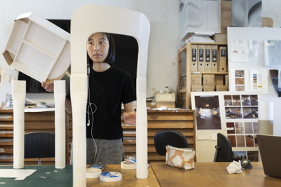 Person arbejder i værksted med en hvid stol/designprototype omgivet af modeller og materialer.