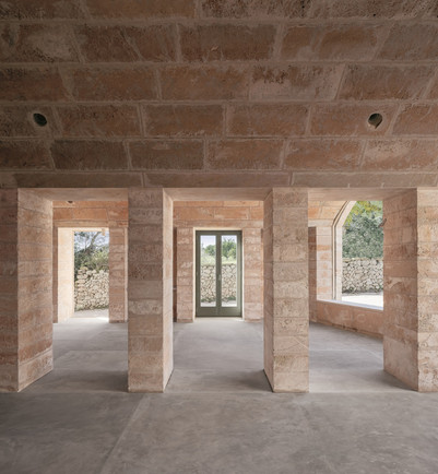 Unfinished interior with light stone columns, arched openings, and a glass door facing a stone-walled courtyard.