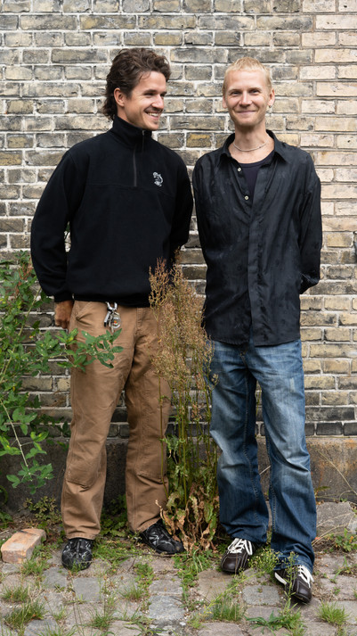 Two men standing together and smiling in front of a brick wall.