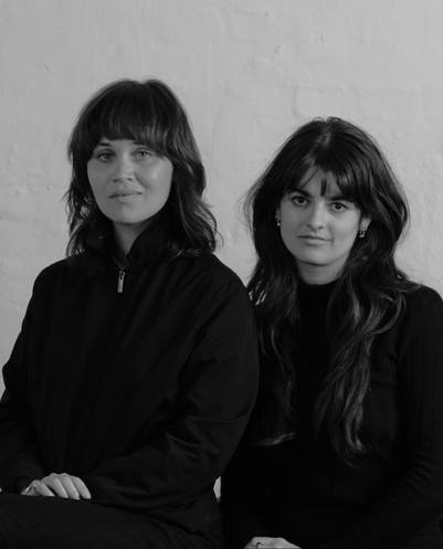 Black-and-white portrait of two dark-haired women in black clothing sitting side by side against a light wall.