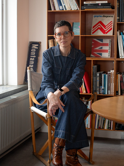Sofie Beier sidder foran en bogreol iført denimtøj, brune støvler og briller.