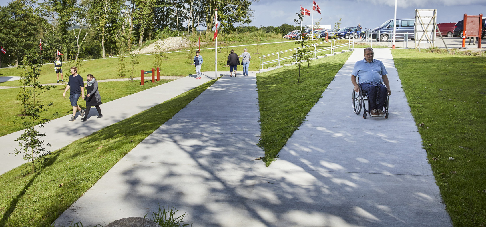 Outdoor path with people, including a person using a wheelchair, surrounded by green areas and Danish flags.