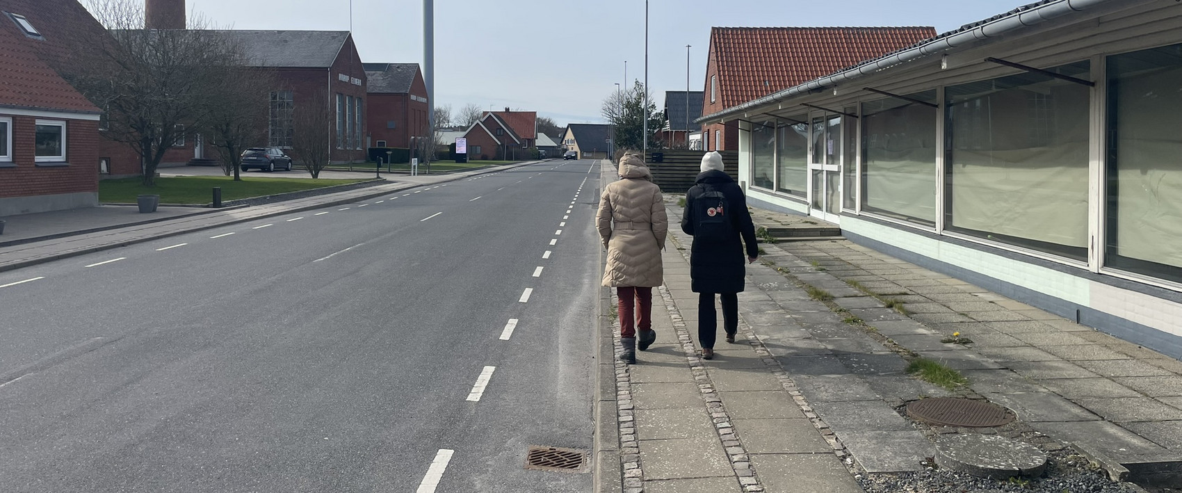 Two people walking along a sidewalk in a small town with low-rise buildings
