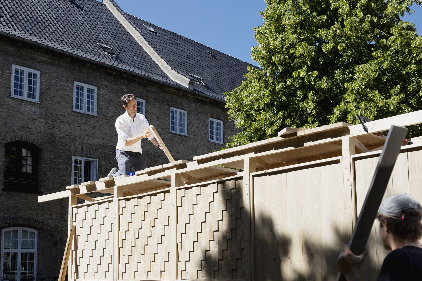 Two men work on a large wooden structure in front of a brick building.